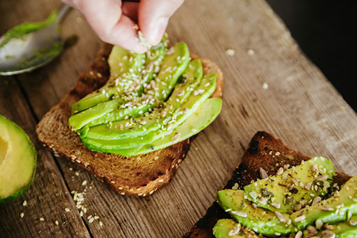 Tafel met halve avocado, toostjes met avocado en iemand die witte zaadjes sprinkelt Tafel met halve avocado, toostjes met avocado en iemand die witte zaadjes sprinkelt