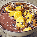 Wit schaaltje met daarin bruine yoghurt versierd met mango en verschillende granen en zaden