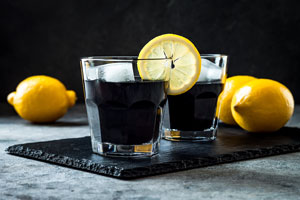 Twee glazen met geactiveerde houtskool limonade op een zwart plakje met daarnaast 2 citroenen