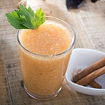 Tafel met daarop een glas met oranje smoothie en een wit schaaltje met 3 kaneelstokjes